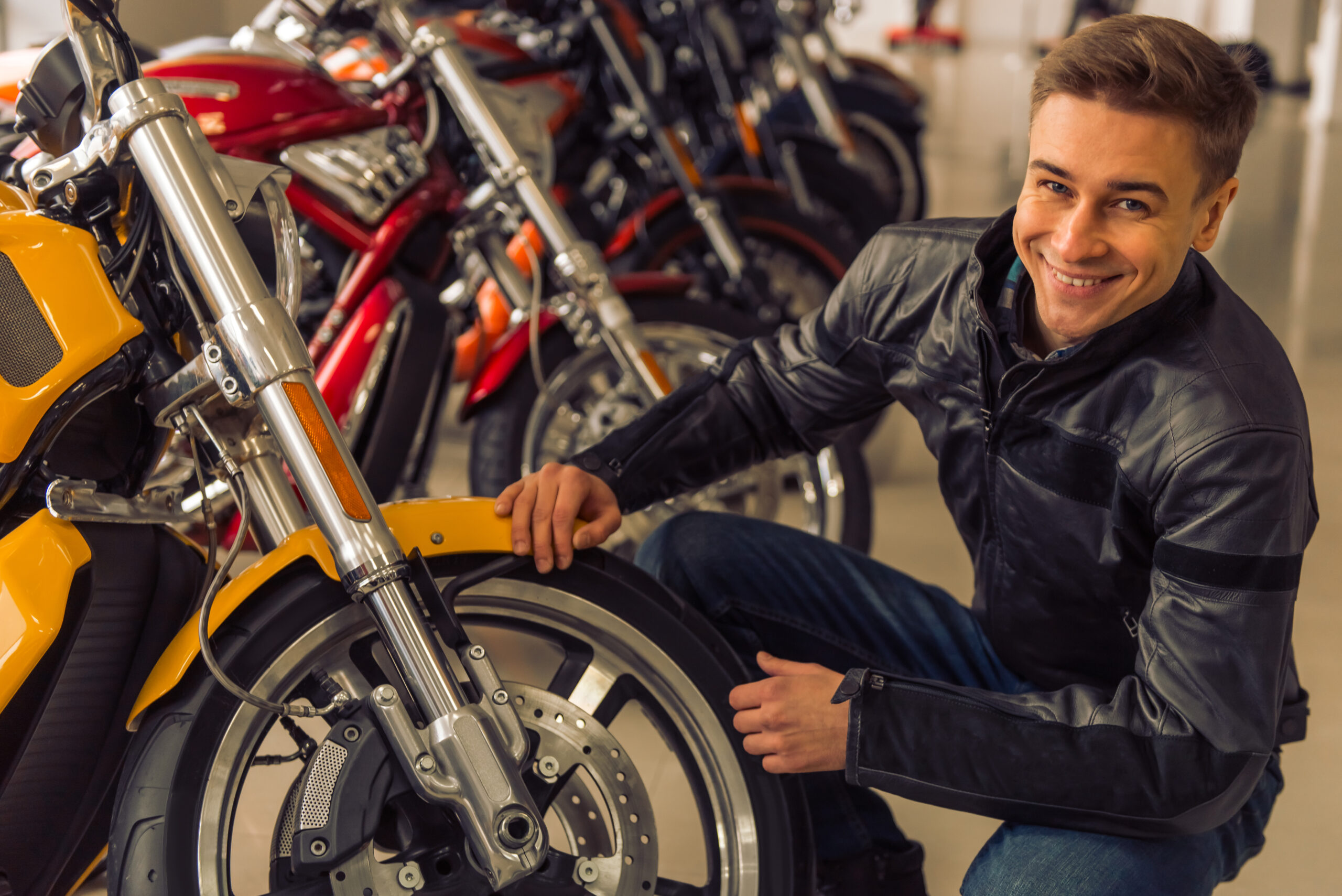 Attractive young blond man in black leather jacket is looking at camera and smiling while examining a motorbike